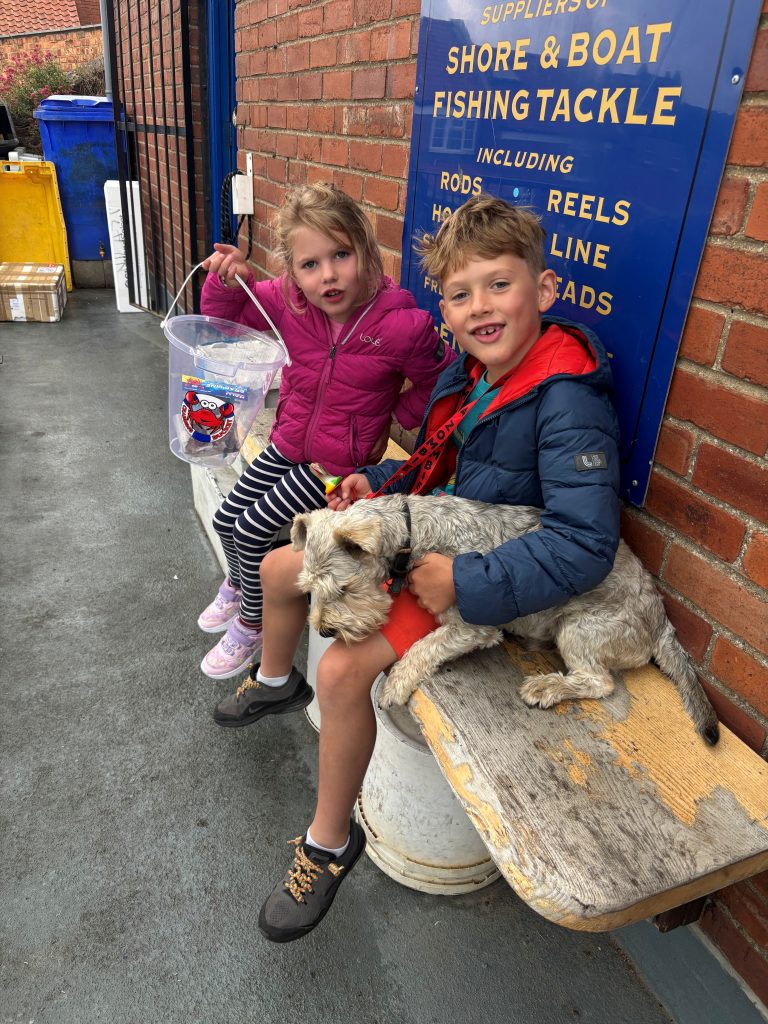 happy boy and girl sitting on bench with dog and pail of crabbing supplies outside of tackle shop