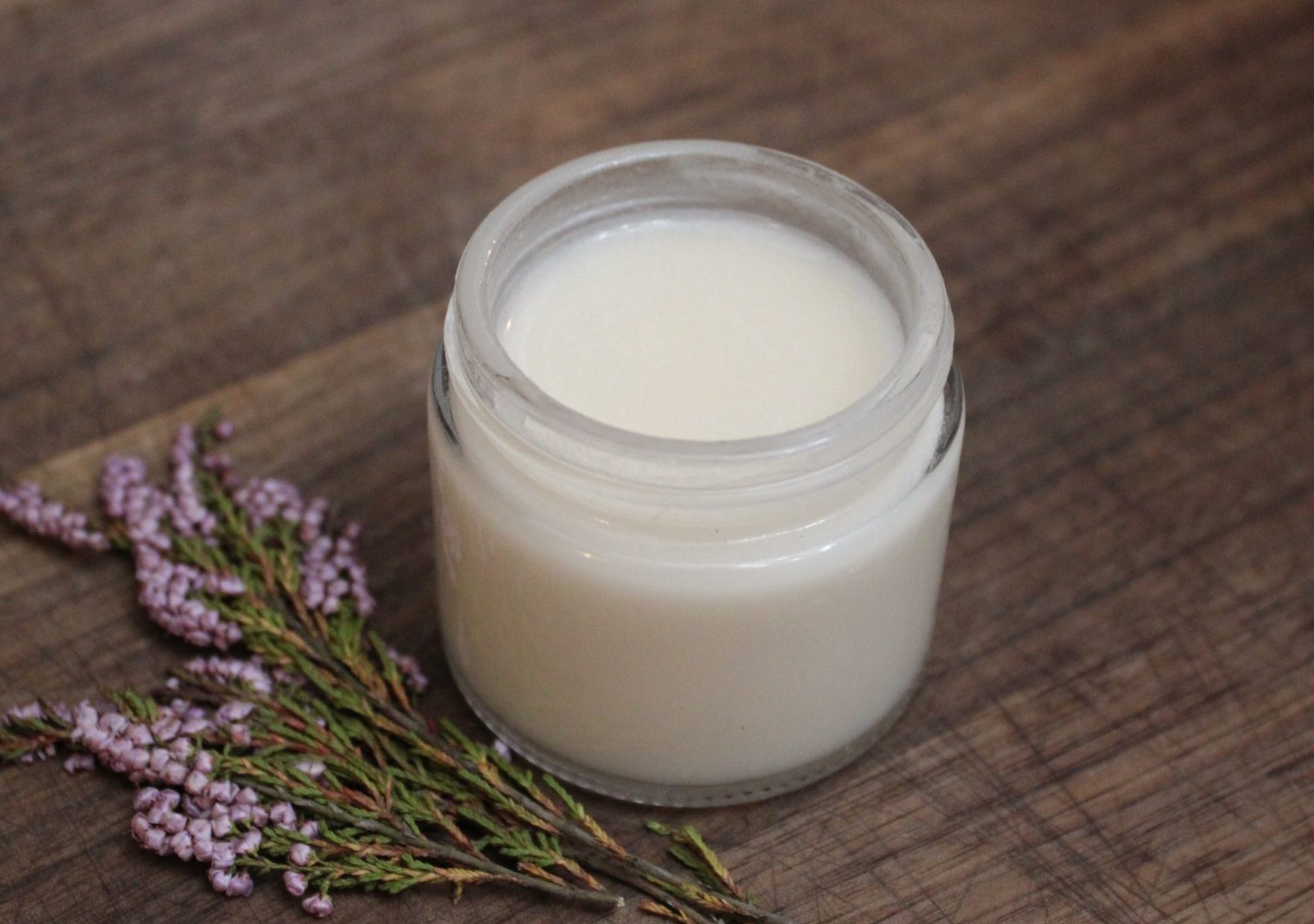 Jar on homemade natural deodorant sitting on top of a dark wood cutting board with dried flowers