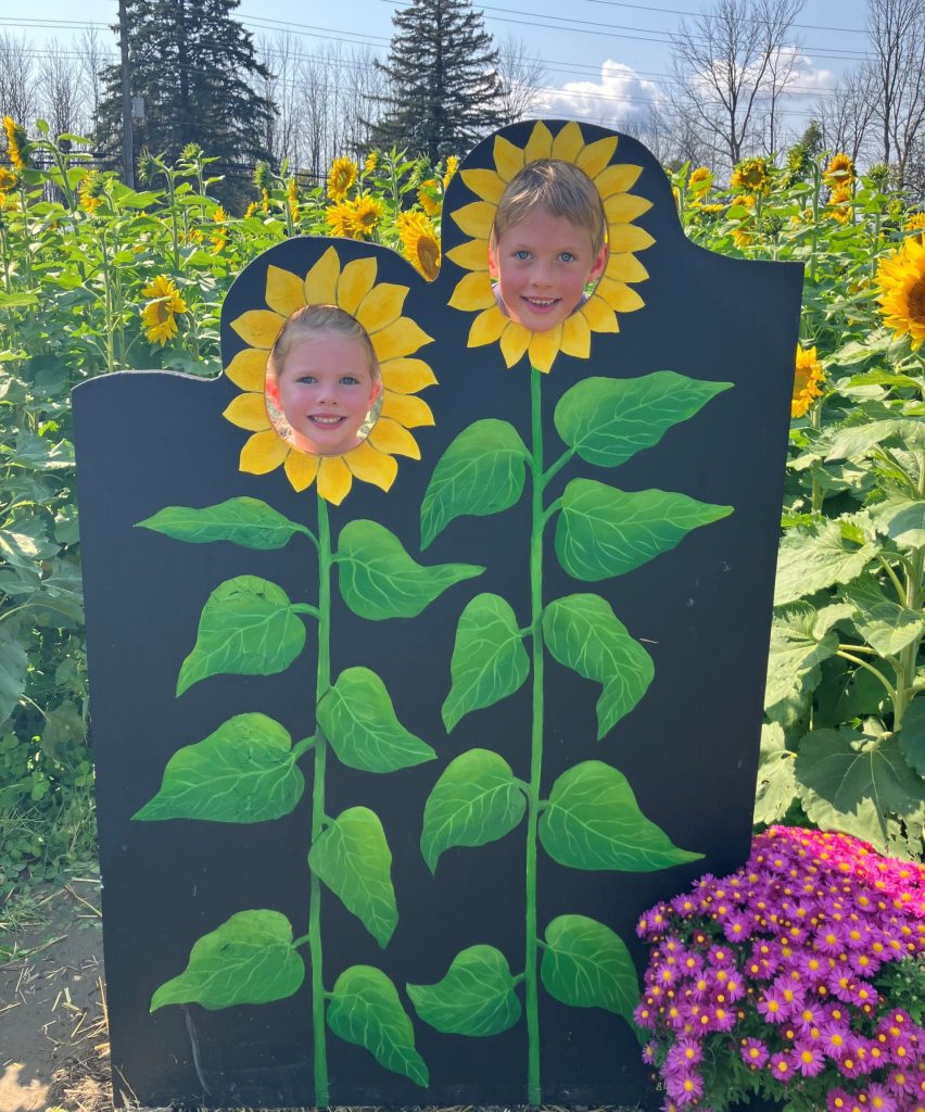 Boy and girl in sunflower patch behind a flower cut out with their faces as sunflowers on a sunny fall day