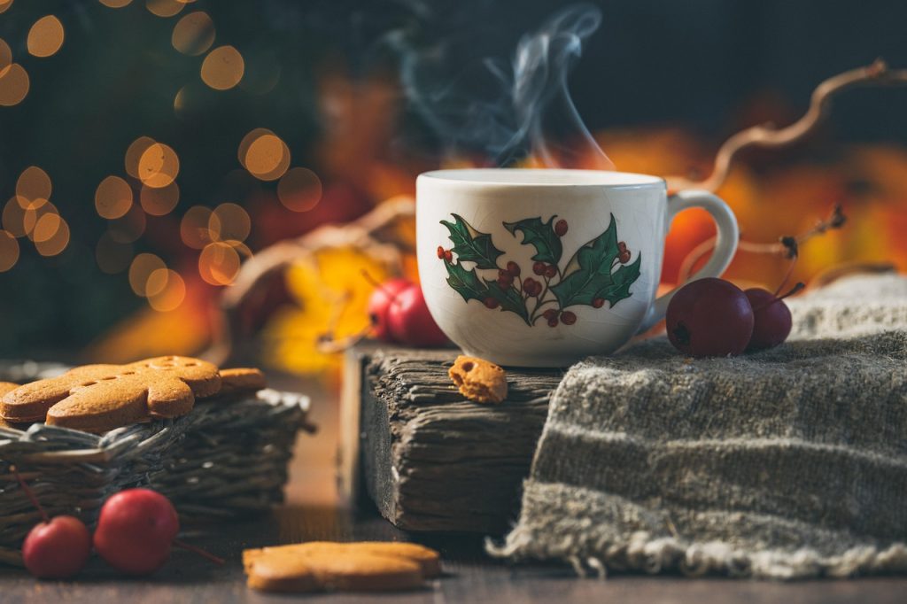 mug of steaming drink sitting on piece of wood with mistletoe and holly printed on mug. Cranberries and gingerbread cookies beside.
