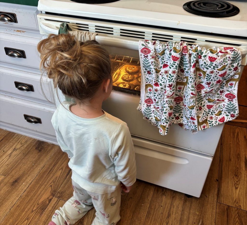 Girl kneeling on floor of kitchen, watching muffins rise through glass on oven door