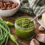 mason jar filled with scape pesto on wooden cutting board with fresh scapes, pecans, garlic, parmesan cheese and a stripes tea towel in background