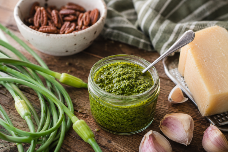 mason jar filled with scape pesto on wooden cutting board with fresh scapes, pecans, garlic, parmesan cheese and a stripes tea towel in background