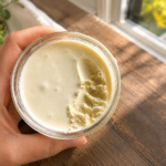 woman holding clear glass jar of light coloured whipped face moisturizer with wood floors in background
