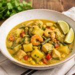 white bowl containing yellow curry seafood chowder on wood countertop with fresh parsley, lime slices, a napkin and a spoon