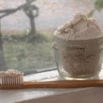 small clear glass jar sitting on window sill containing gritty off white natural toothpaste and bamboo toothbrush in front of it