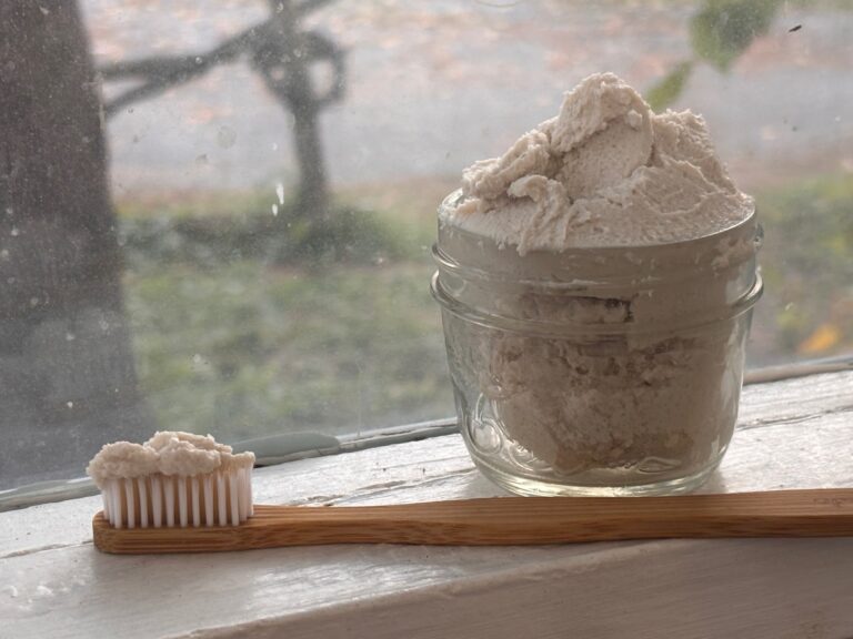 small clear glass jar sitting on window sill containing gritty off white natural toothpaste and bamboo toothbrush in front of it